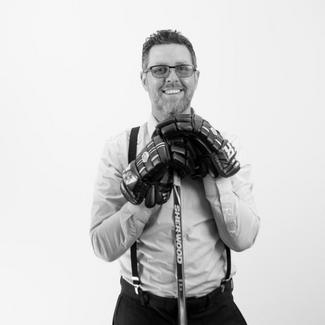 A smilling man in wearing a dress shirt, pants and hockey gloves.